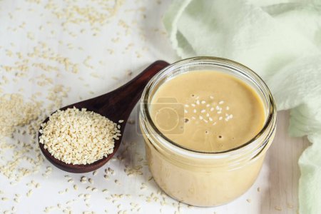 Photo for Top view of mason jar filled with tahini. Wooden spoon overflowing with sesame seeds. Top view. - Royalty Free Image