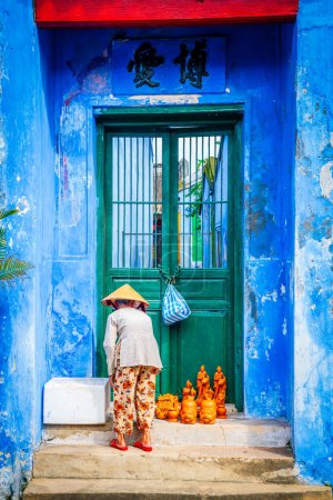 Photo for Hoy An, Vietnam, November 20, 2022: An elderly lady is setting up a display of hand-crafted statues on the steps of a temple in Hoy An, Vietnam - Royalty Free Image