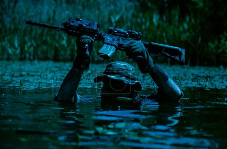 A soldier moves in the heart of a marsh, submerged in swampy waters with only arms and rifle visible, extreme conditions of concealed tactical combat night operation