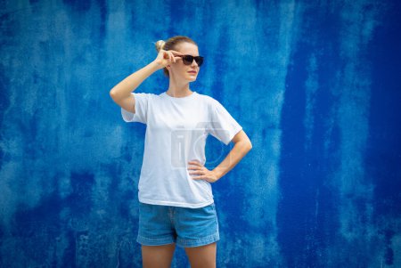 Téléchargez les photos : Modèle féminin portant un t-shirt blanc blanc sur le fond d'un mur bleu - en image libre de droit