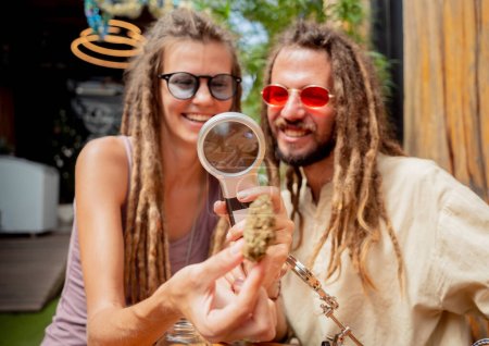 Foto de Estilo hippie pareja examina bajo una lupa las articulaciones y brotes de marihuana medicinal. - Imagen libre de derechos
