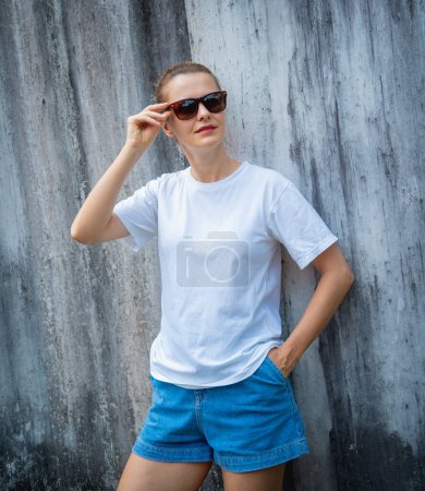 Téléchargez les photos : Modèle féminin portant un t-shirt blanc blanc sur le fond d'un mur gris rayé - en image libre de droit