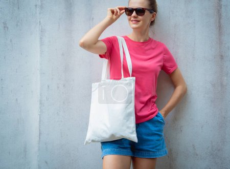 Téléchargez les photos : Modèle féminin portant un t-shirt blanc rose sur le fond d'un mur gris - en image libre de droit