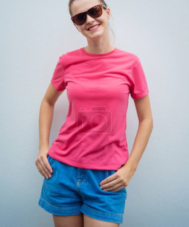 Téléchargez les photos : Modèle féminin portant un t-shirt blanc rose sur le fond d'un mur gris - en image libre de droit