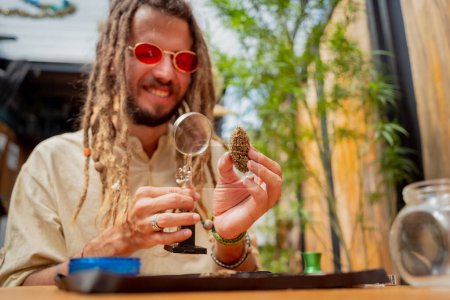 Photo for Hippie style man examines under a magnifying glass the joints and buds of medical marijuana. - Royalty Free Image