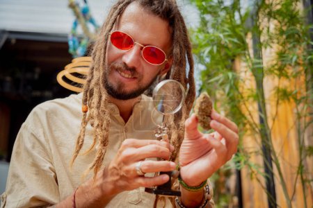 Foto de Hippie hombre estilo examina bajo una lupa las articulaciones y brotes de marihuana medicinal. - Imagen libre de derechos