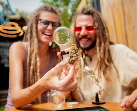Foto de Estilo hippie pareja examina bajo una lupa las articulaciones y brotes de marihuana medicinal. - Imagen libre de derechos