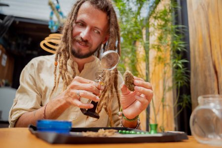 Foto de Hippie hombre estilo examina bajo una lupa las articulaciones y brotes de marihuana medicinal. - Imagen libre de derechos