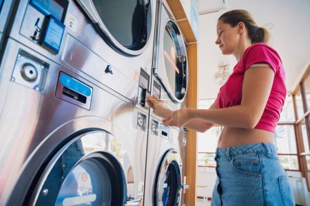 Photo for Young woman putting coin in washing machine at laundry. - Royalty Free Image