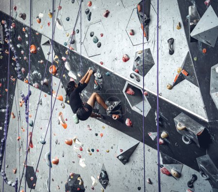 Téléchargez les photos : Jeune homme sur mur d'escalade artificiel avec poignées et cordes colorées. - en image libre de droit