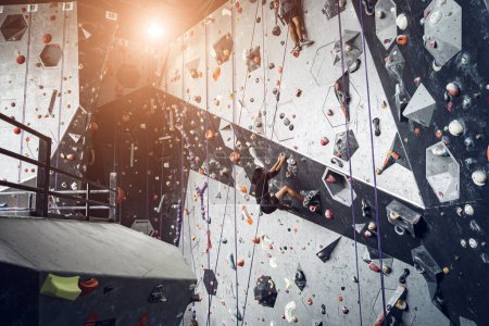 Téléchargez les photos : Jeune homme sur mur d'escalade artificiel avec poignées et cordes colorées. - en image libre de droit