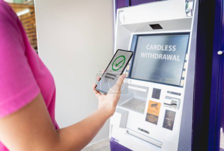 Foto de Mujer joven usando el teléfono inteligente para retirar sin tarjeta el dinero cerca del cajero automático. - Imagen libre de derechos