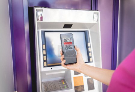 Téléchargez les photos : Jeune femme utilisant le téléphone intelligent pour retirer sans carte l'argent près du distributeur automatique de billets. - en image libre de droit