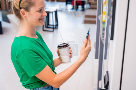 Photo for Young woman paying for coffee at vending machine using contactless method of payment. - Royalty Free Image
