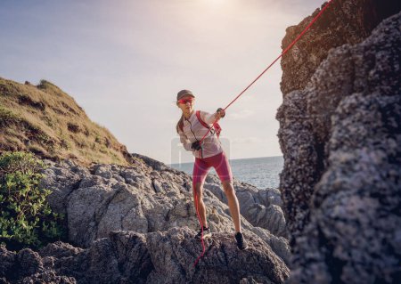 Photo for Young woman with a rope climbs to the top in the mountains near the ocean. - Royalty Free Image