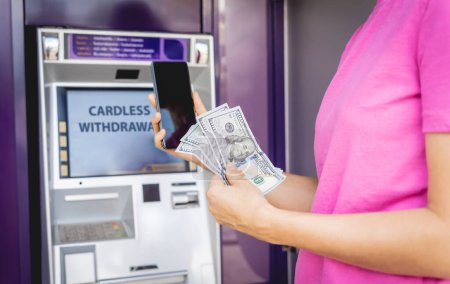 Foto de Mujer joven usando el teléfono inteligente para retirar sin tarjeta el dinero cerca del cajero automático. - Imagen libre de derechos