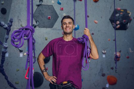 Téléchargez les photos : Un grimpeur mâle fort contre un mur artificiel avec des poignées et des cordes colorées - en image libre de droit