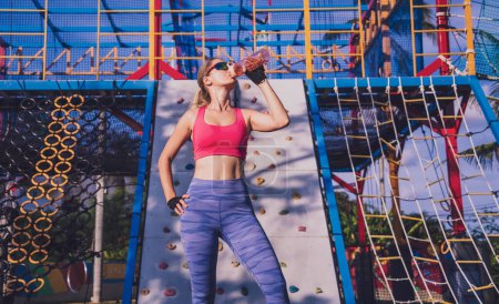 Téléchargez les photos : Jeune femme sportive qui boit de l'eau après s'être entraînée et avoir escaladé le rocher artificiel dans un camp d'entraînement - en image libre de droit