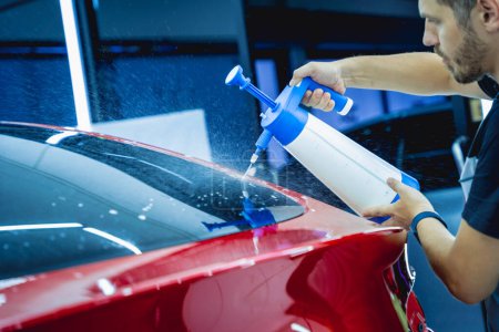 Téléchargez les photos : Travailleur en service automobile pulvérise de l'eau sur la voiture avant d'appliquer un film protecteur - en image libre de droit
