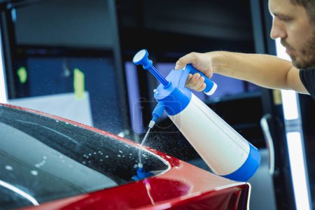 Téléchargez les photos : Travailleur en service automobile pulvérise de l'eau sur la voiture avant d'appliquer un film protecteur - en image libre de droit