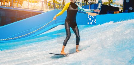 Téléchargez les photos : Belle jeune femme surfant sur un simulateur de vagues dans un parc d'attractions aquatiques. - en image libre de droit