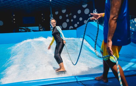 Foto de Hermosa joven surfeando con entrenador en un simulador de olas en un parque de atracciones acuático. - Imagen libre de derechos