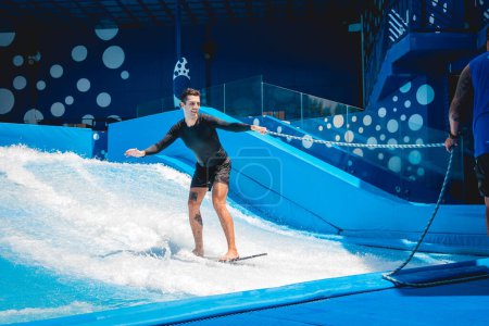Téléchargez les photos : Jeune homme surfant avec un entraîneur sur un simulateur d'onde dans un parc d'attractions aquatiques. - en image libre de droit
