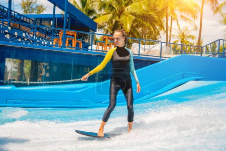 Téléchargez les photos : Belle jeune femme surfant sur un simulateur de vagues dans un parc d'attractions aquatiques. - en image libre de droit