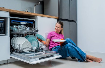Téléchargez les photos : Jeune femme assise par terre près de la machine à laver le vaisselle. - en image libre de droit