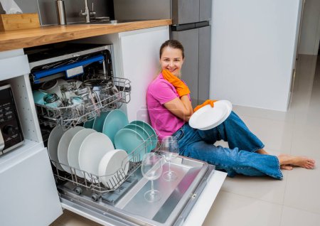 Foto de Mujer joven sentada en el suelo cerca de la máquina lavavajillas. - Imagen libre de derechos