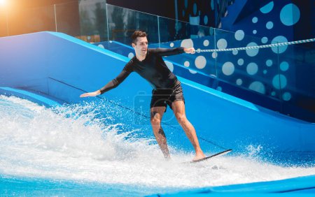 Téléchargez les photos : Jeune homme surfant avec un entraîneur sur un simulateur d'onde dans un parc d'attractions aquatiques. - en image libre de droit