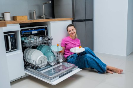 Photo for Young woman sitting on the floor near the dishwasher machine. - Royalty Free Image