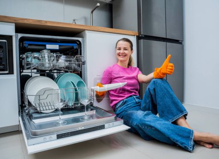 Téléchargez les photos : Jeune femme assise par terre près de la machine à laver le vaisselle. - en image libre de droit