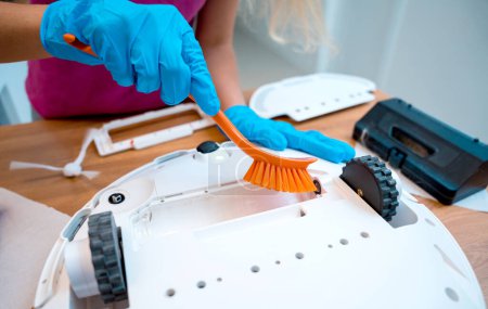 Téléchargez les photos : Une jeune femme nettoie un robot aspirateur de la saleté après le nettoyage - en image libre de droit