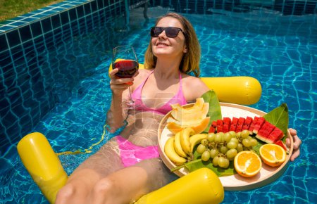 Téléchargez les photos : Belle femme couchée sur hamac flottant dans la piscine avec vin et plateau de fruits. - en image libre de droit