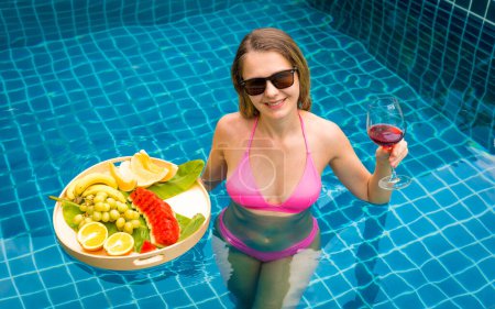 Téléchargez les photos : Belle femme dans la piscine avec verre de vin et plateau flottant de fruits. - en image libre de droit