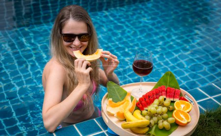 Téléchargez les photos : Belle femme dans la piscine avec verre de vin et plateau flottant de fruits. - en image libre de droit