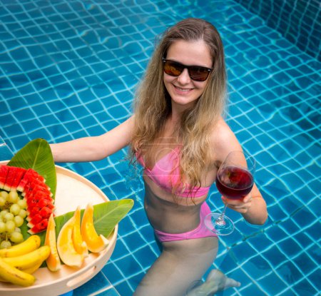 Foto de Hermosa mujer en la piscina con copa de vino y bandeja flotante de frutas. - Imagen libre de derechos