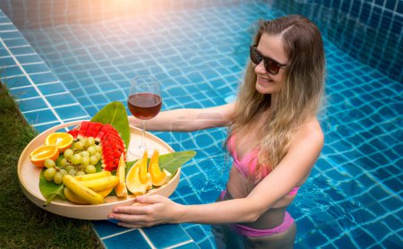 Foto de Hermosa mujer en la piscina con copa de vino y bandeja flotante de frutas. - Imagen libre de derechos
