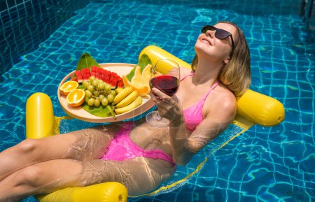 Photo for Beautiful woman lying on floating hammock in the swimming pool with wine and tray of fruits. - Royalty Free Image