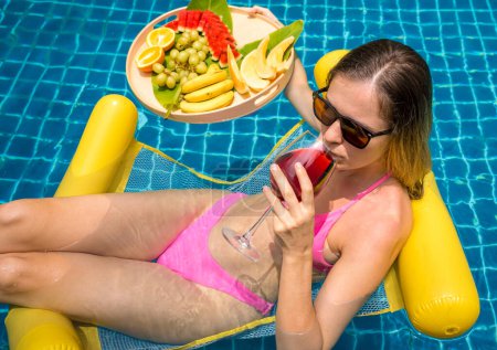 Téléchargez les photos : Belle femme couchée sur hamac flottant dans la piscine avec vin et plateau de fruits. - en image libre de droit