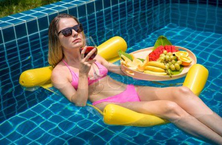 Photo for Beautiful woman lying on floating hammock in the swimming pool with wine and tray of fruits. - Royalty Free Image