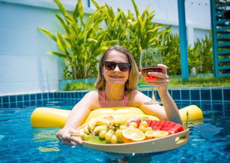 Foto de Hermosa mujer acostada en hamaca flotante en la piscina con vino y bandeja de frutas. - Imagen libre de derechos
