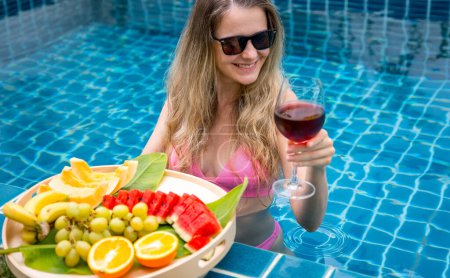Photo for Beautiful woman in the swimming pool with glass of wine and floating tray of fruits. - Royalty Free Image