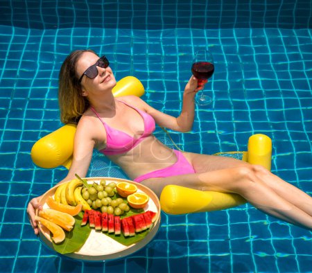 Foto de Hermosa mujer acostada en hamaca flotante en la piscina con vino y bandeja de frutas. - Imagen libre de derechos