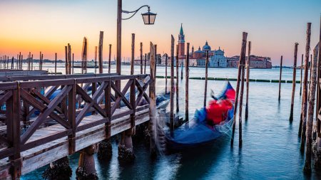 Foto de Amanecer sereno sobre Venecia con góndola borrosa en movimiento por muelles de madera amarrados por la plaza de San Marcos con la iglesia de San Giorgio di Maggiore en el fondo, Venecia, Italia - Imagen libre de derechos