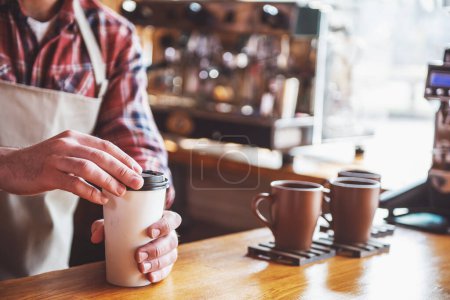 Foto de Imagen recortada de barista guapo en delantal sosteniendo una taza de café mientras está de pie en el mostrador del bar en la cafetería - Imagen libre de derechos