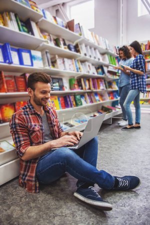 Foto de Estudiante guapo está utilizando un ordenador portátil y sonriendo mientras está sentado en la librería, en el fondo las niñas están eligiendo libros - Imagen libre de derechos