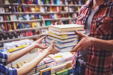 Photo for Cropped image of young man and woman holding a pile of books while standing in the book shop - Royalty Free Image