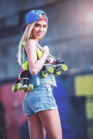Photo for Side view of beautiful blonde girl in cap looking at camera and smiling while standing with her rollers in skate park - Royalty Free Image